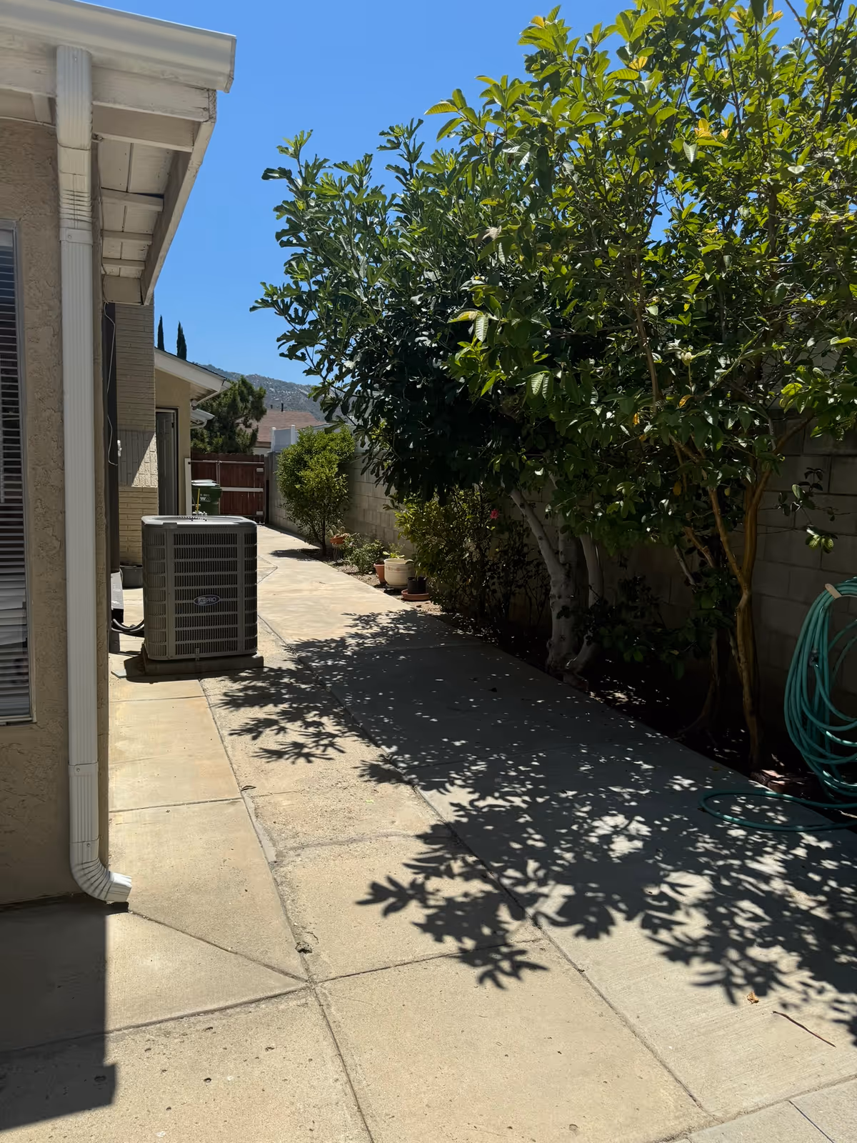 Side yard concrete walkway beside a house with an air conditioning unit, trees casting shadows, potted plants and a garden hose.