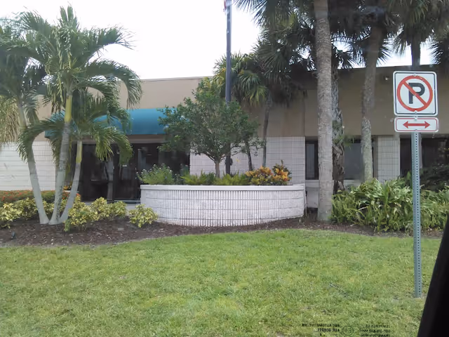 Entrance of a low-rise nursing center with palm trees, a raised planter and a No Parking sign on a grassy lawn.