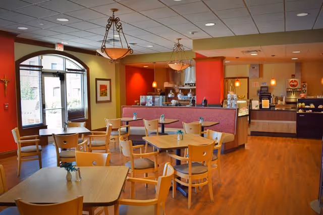 A bright and clean dining area with wooden tables and chairs arranged neatly. The room features warm red and yellow walls, pendant lighting, and a counter area with coffee machines and food displays. Large windows and a glass door allow natural light to enter the space.