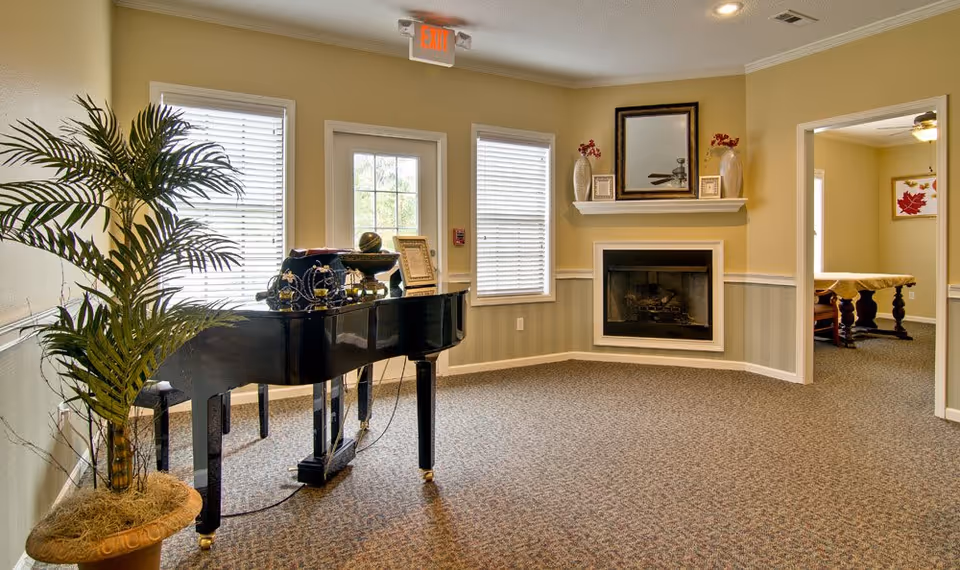 A cozy interior room with a black grand piano adorned with decorative items, a potted plant to the left, two windows with blinds, and a door with an exit sign above it. On the right side, there is a fireplace with a mantel holding vases and framed pictures, and an open doorway leading to another room with a table and framed artwork on the wall.