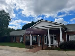 Exterior view of Woodland Place Senior Living building with a covered entrance, white columns, and a rocking chair on the porch under a partly cloudy sky.