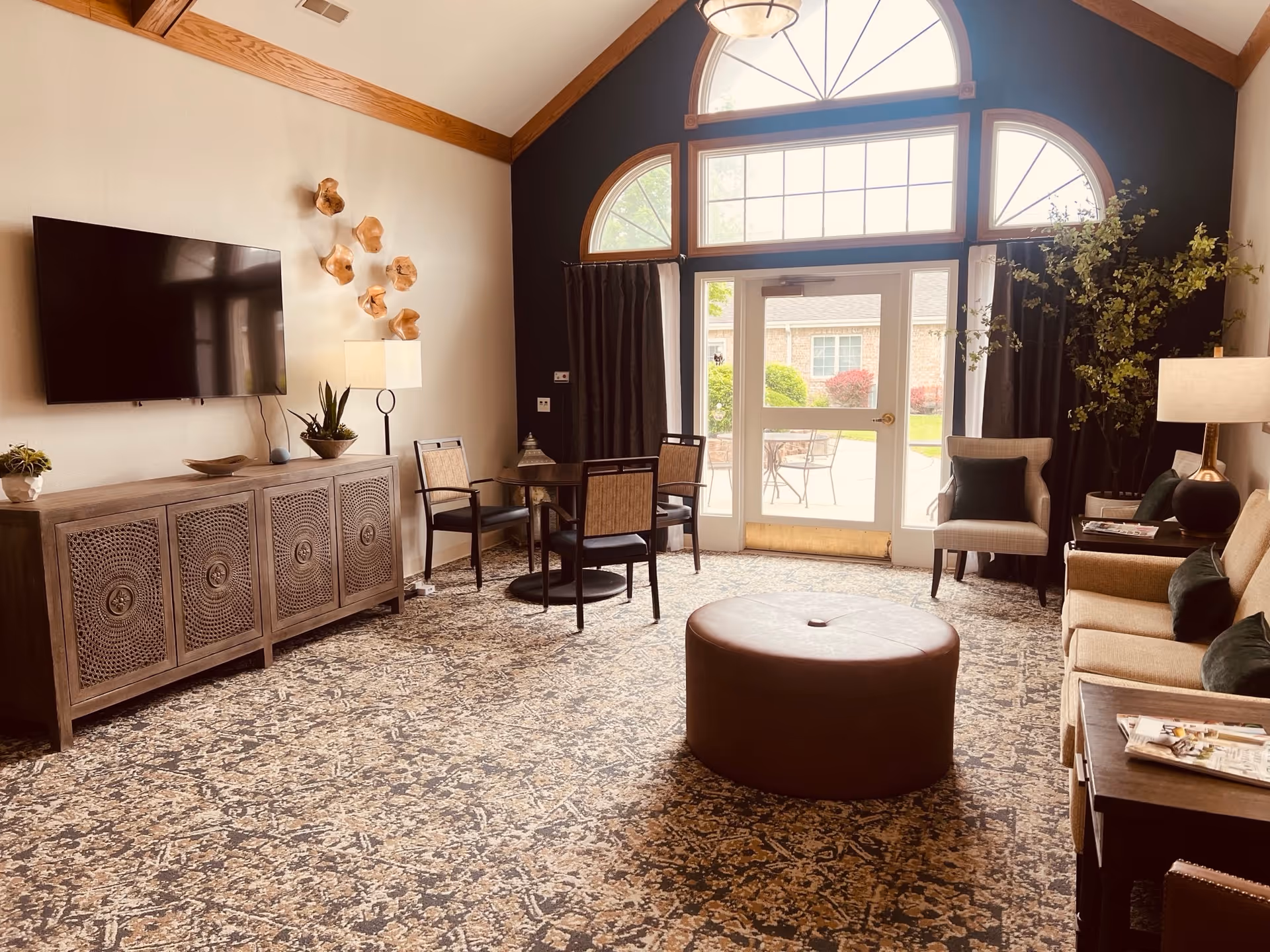 Well-lit lounge with seating, a wall-mounted TV, round ottoman, and large arched windows/door opening to an outdoor patio.