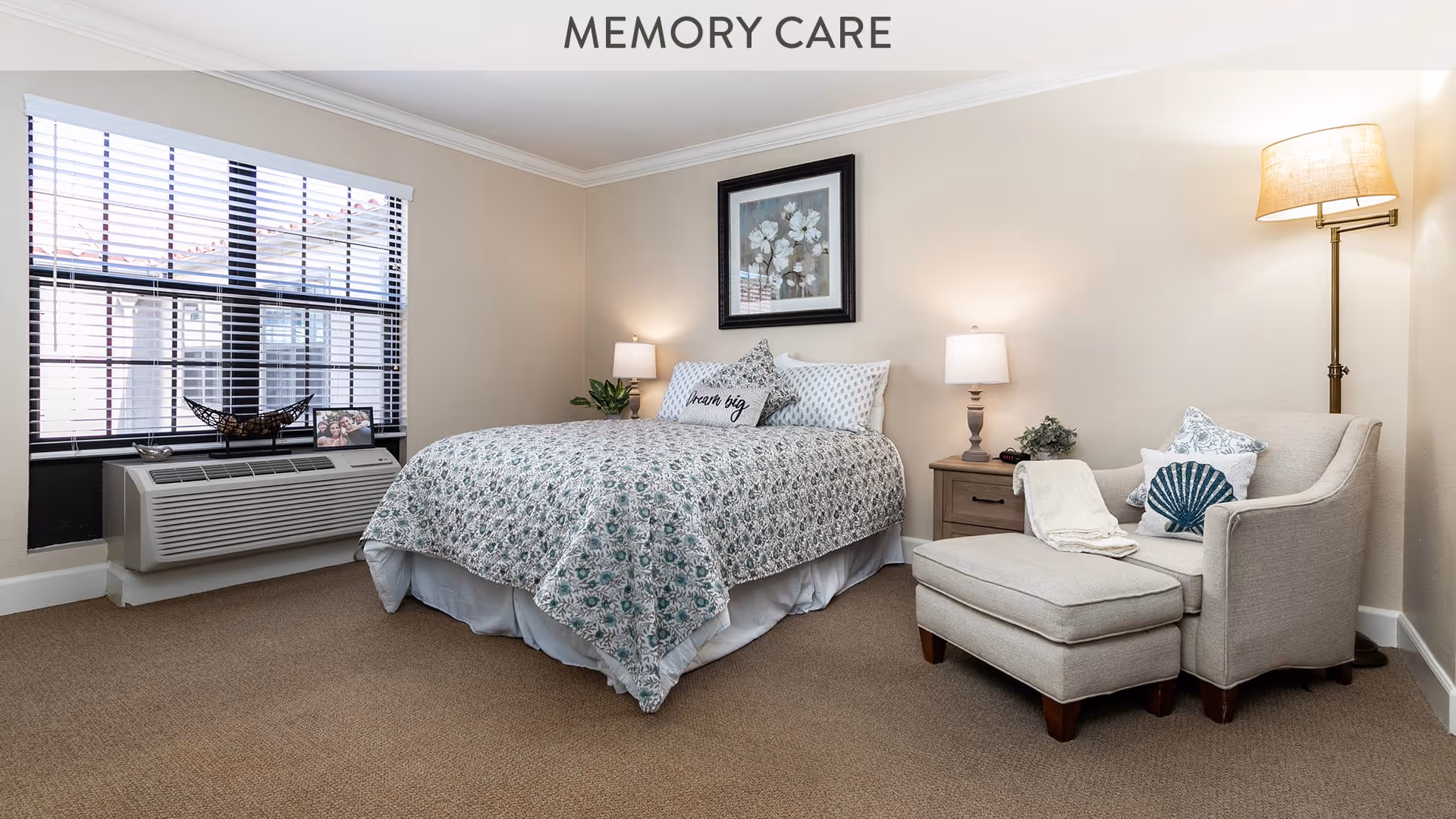 A cozy bedroom in a memory care facility featuring a bed with floral bedding and pillows, a nightstand with a lamp and plant, a comfortable armchair with an ottoman, a floor lamp, and a large window with blinds.