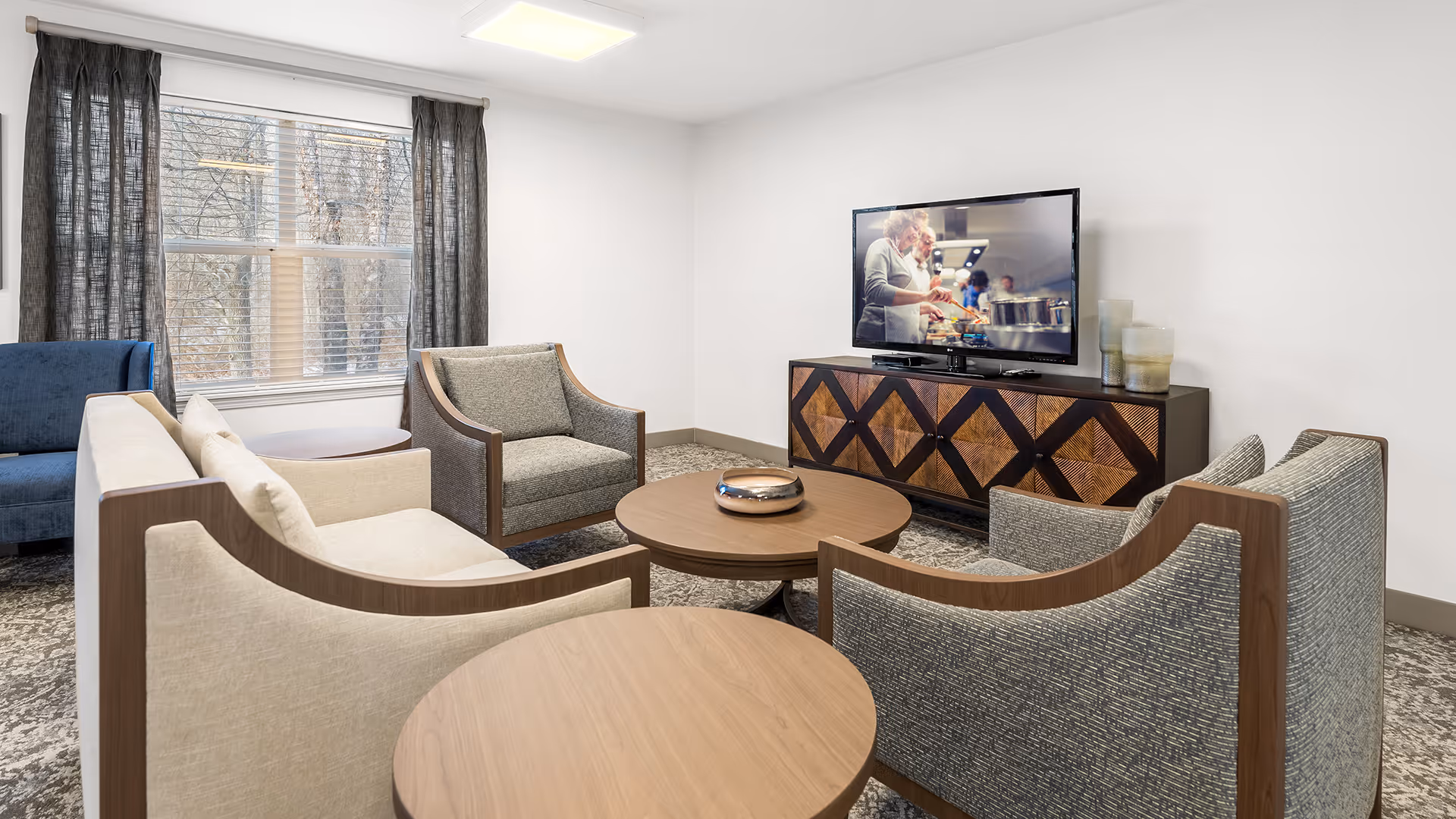 A cozy living room area with a large window covered by dark curtains. The room features a beige sofa, two gray armchairs, a blue chair, and two round wooden coffee tables. A flat-screen TV is mounted on a wooden cabinet with a geometric pattern, displaying a cooking show.