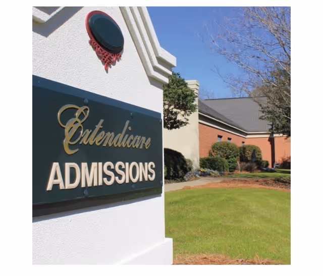 Outdoor view of a sign that reads 'Extendicare ADMISSIONS' mounted on a white structure with a building and landscaped greenery in the background under a clear blue sky.