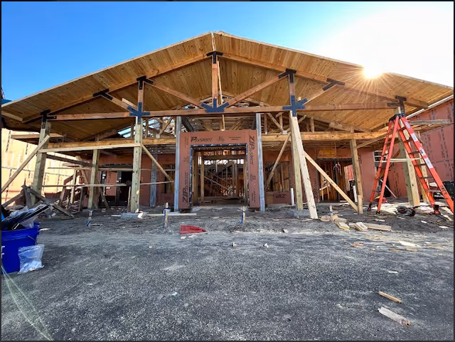A building under construction with wooden framing and a pitched roof. The structure is supported by temporary wooden braces, and construction materials and tools are scattered around the site. The sun is shining brightly in the clear blue sky.