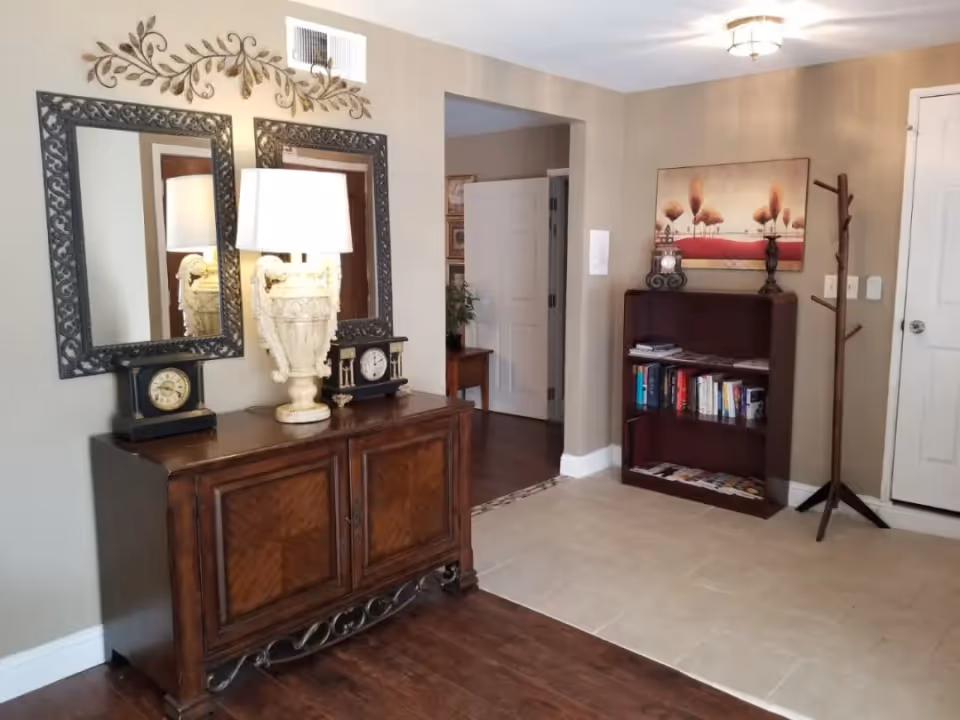 A foyer-style interior with a wooden sideboard topped by a lamp and clocks under two mirrors, and a bookshelf and coat rack against neutral walls.