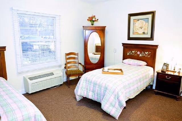 Bright, simply furnished bedroom with two twin beds (one partly visible), wooden headboards, a mirrored wardrobe, chair, and nightstand under a window.
