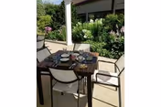 Patio dining table set for four with plates and cups on a sunny terrace next to a garden and building windows.