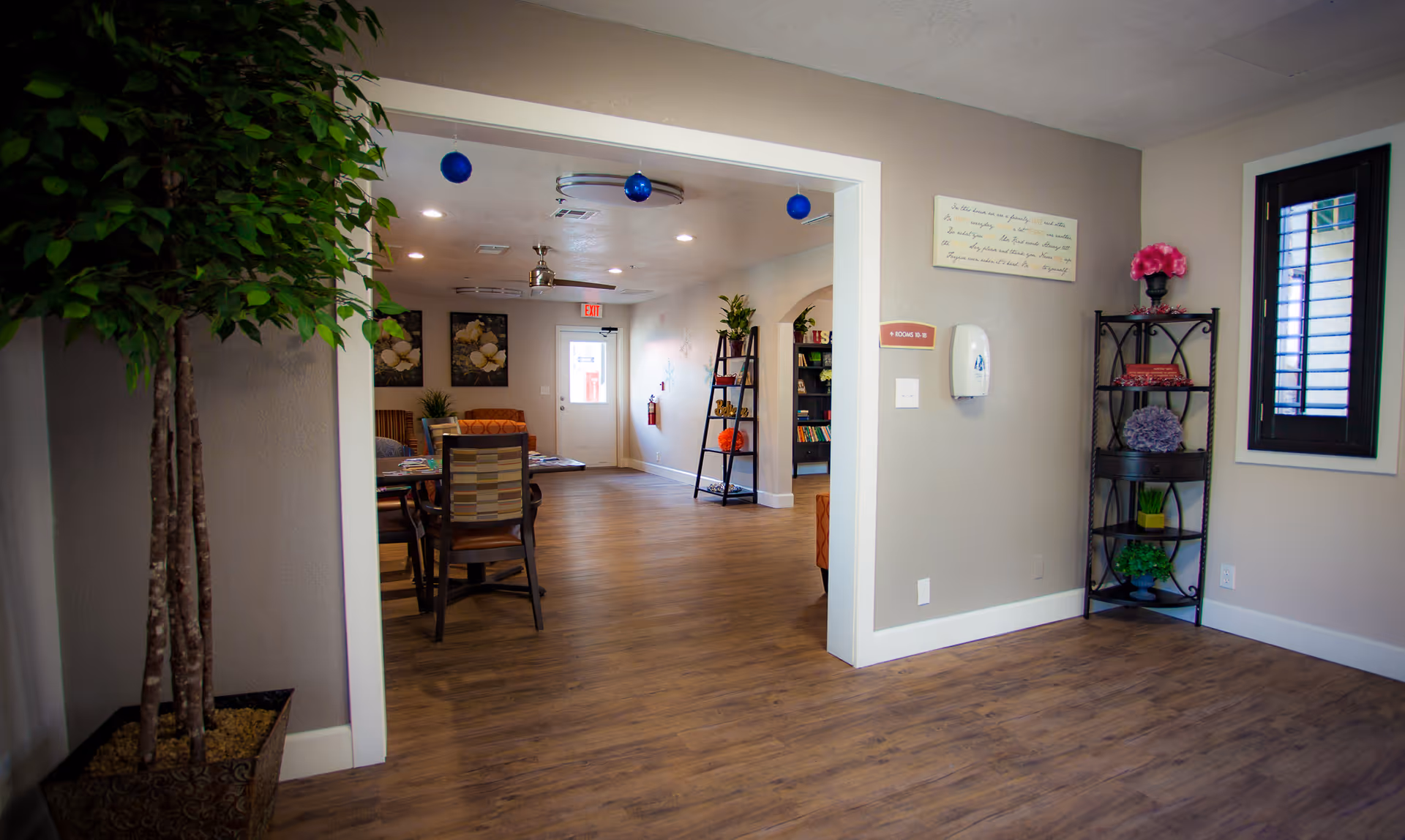 Interior view of a common area in a senior living facility with wooden flooring, a potted plant on the left, a black metal shelf with decorative items on the right, and a dining table with chairs in the background. The walls are light-colored with framed artwork and a sign indicating rooms 10-18. Blue hanging ornaments are visible from the ceiling.