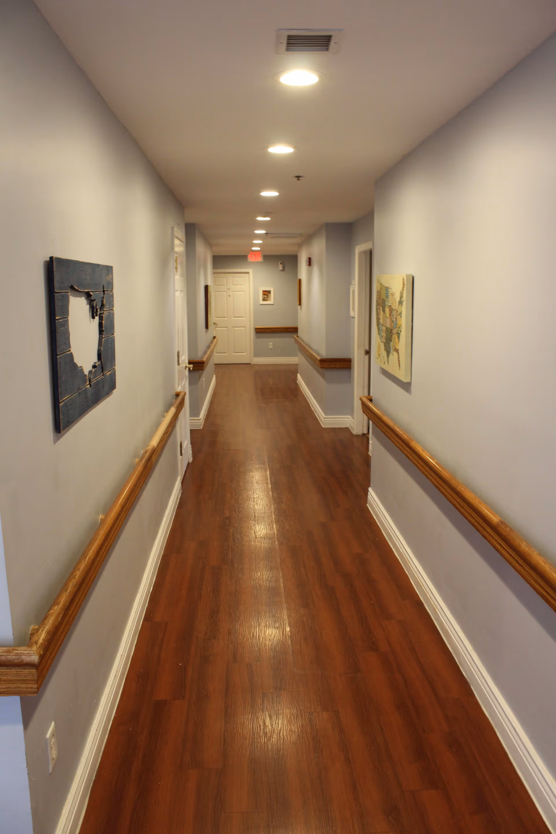A long, well-lit hallway with wooden flooring and light gray walls. Handrails run along both sides of the hallway, which is decorated with framed artwork. Several doors line the hallway, and ceiling lights illuminate the space.