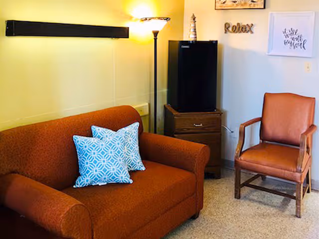A cozy sitting area with an orange couch adorned with two blue patterned pillows, a wooden armchair with brown cushions, a small wooden cabinet with a black mini fridge on top, a tall floor lamp, and wall decorations including a 'Relax' sign and framed artwork.