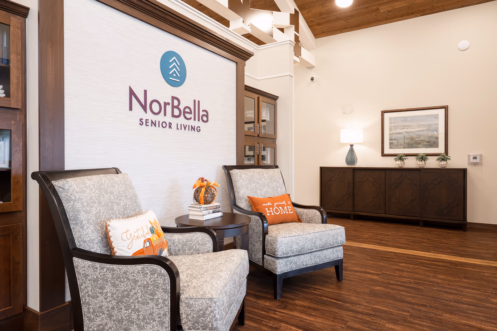 A cozy seating area in NorBella Senior Living featuring two patterned armchairs with decorative pillows, a small round table with books and a pumpkin decoration, a wooden cabinet with a lamp and framed artwork on the wall, and a NorBella Senior Living logo displayed prominently on a textured wall panel.