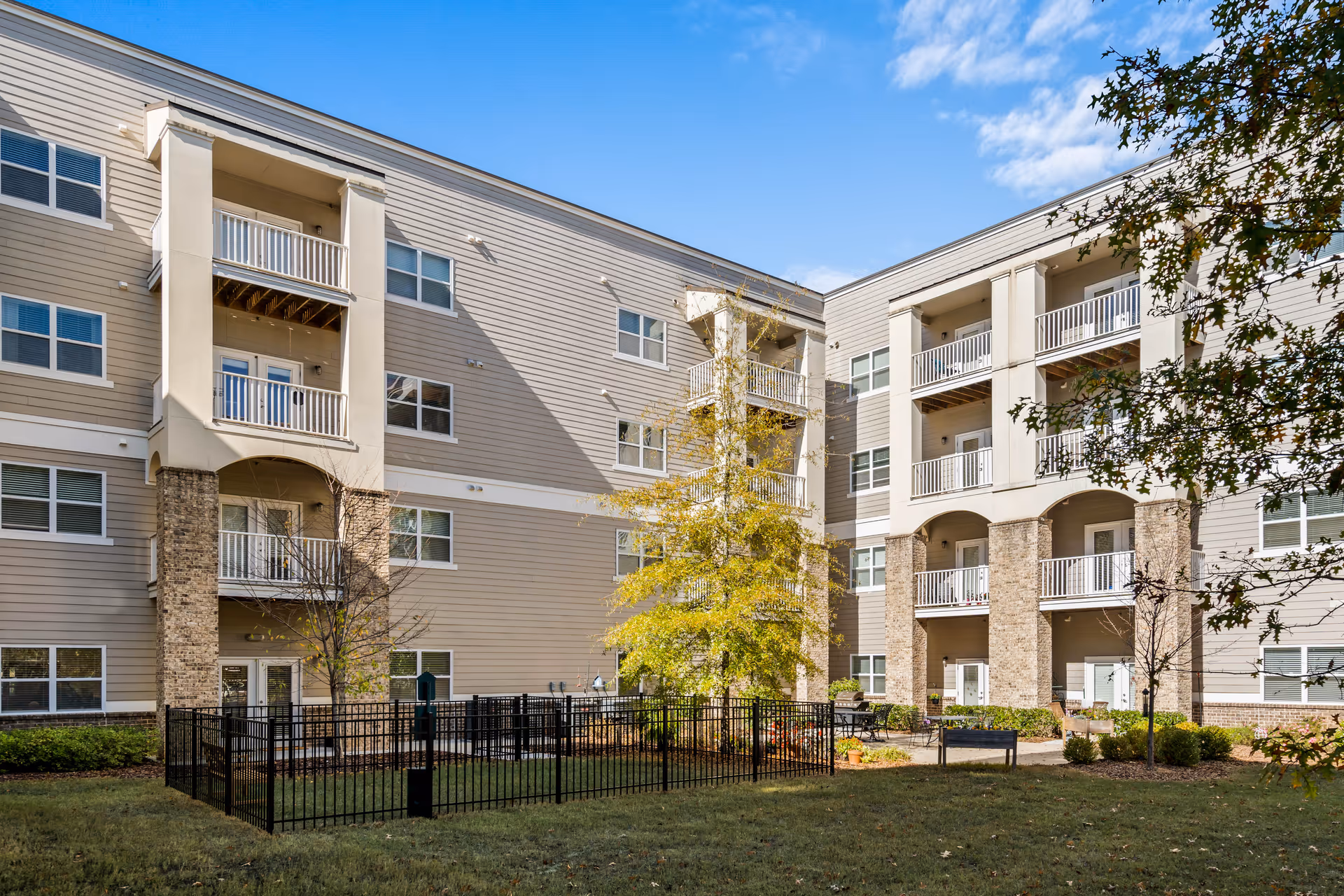 Exterior view of a senior living facility building with multiple balconies, beige siding, and brick accents. There is a fenced grassy area with a tree and some shrubs in the courtyard under a clear blue sky.