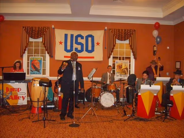 A musical band performing indoors in front of a USO banner. The band includes a singer holding a microphone, a keyboard player, a drummer, and several musicians playing brass instruments. The room has orange walls, two windows with brown curtains, and vintage war bond posters on the walls.