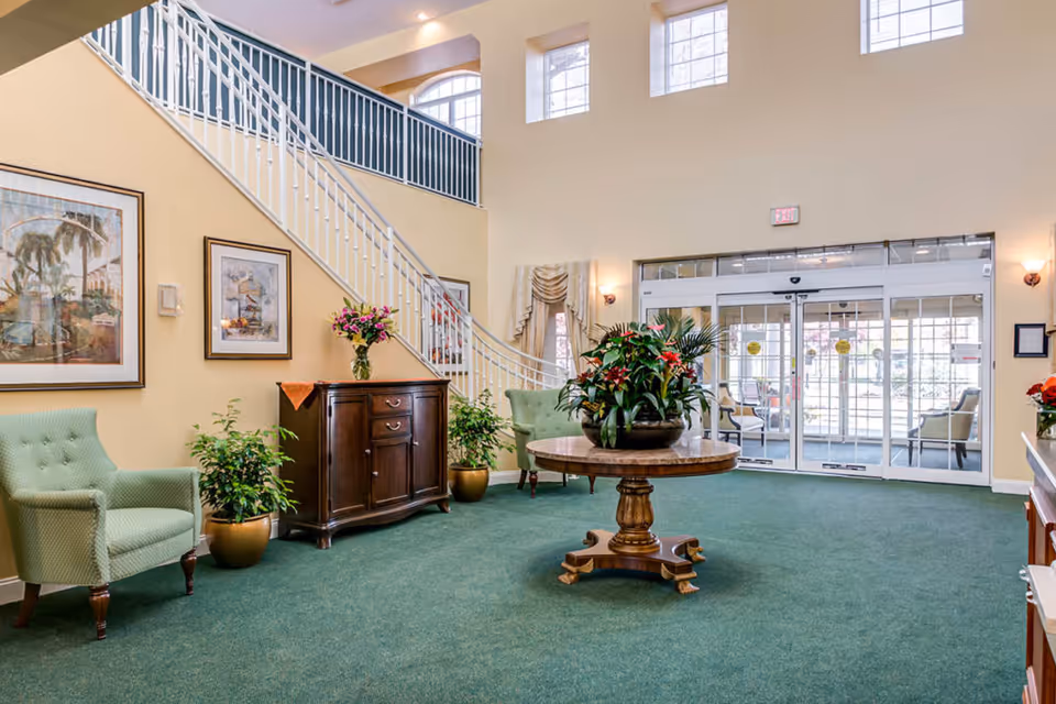 Spacious and well-lit lobby area with green carpet, a round wooden table with a large floral arrangement in the center, two green upholstered armchairs, a wooden cabinet with a vase of flowers, potted plants, framed artwork on the walls, a staircase with white railings, and large glass double doors leading outside.