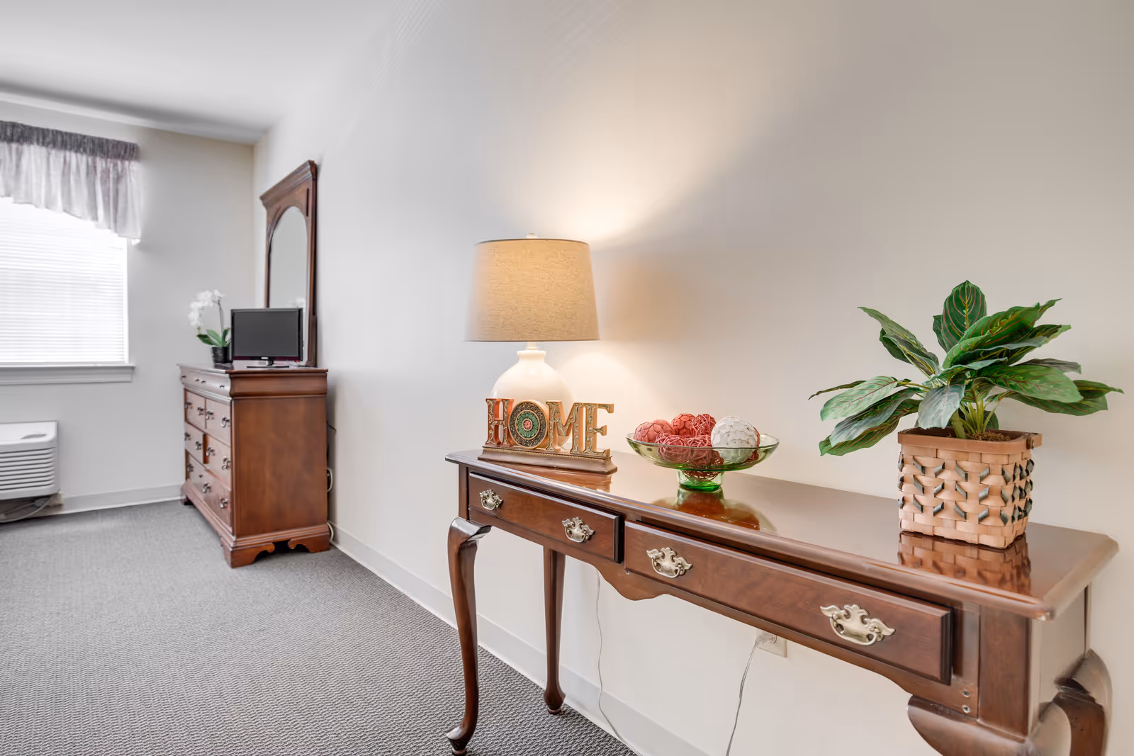 A furnished senior living bedroom area with a wooden console table holding a lamp, decorative bowl and potted plant, and a dresser with mirror and small TV near a window.