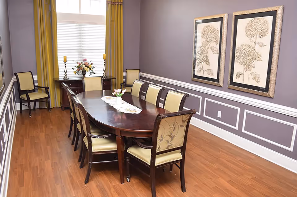 A dining room with a long dark wooden table surrounded by eight chairs with light-colored cushions. The room has purple walls with white trim, two framed floral artworks on one wall, and a window with yellow curtains. A sideboard beneath the window holds two candlesticks and a floral arrangement.