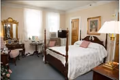 A cozy bedroom featuring a wooden four-poster bed with white bedding and pink pillows. The room includes a vintage vanity with a mirror and cushioned stool, a small round table with two chairs near the window, a television on a stand, and framed artwork on the walls. The room has light-colored walls and carpeted flooring.