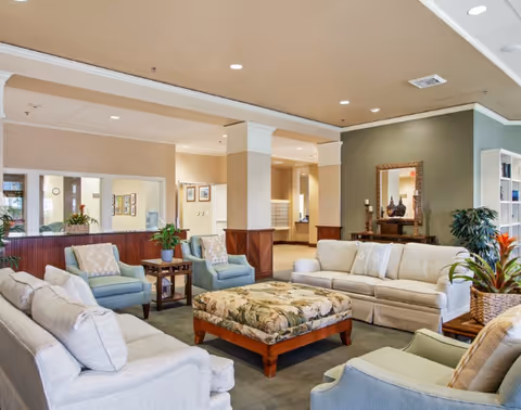 A spacious and well-lit living room area with beige and light blue upholstered sofas and armchairs arranged around a large floral-patterned ottoman. The room features a green accent wall with a decorative mirror and a console table with vases, potted plants, and soft overhead lighting. The background shows an open hallway and mailboxes, suggesting a communal space in a senior living facility.