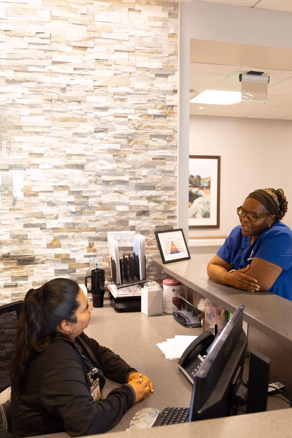 Two women engaged in conversation at a reception desk in a healthcare facility. One woman is seated behind the desk with a computer and office supplies, while the other woman, dressed in blue scrubs, leans on the counter. The background features a textured stone wall and framed artwork.
