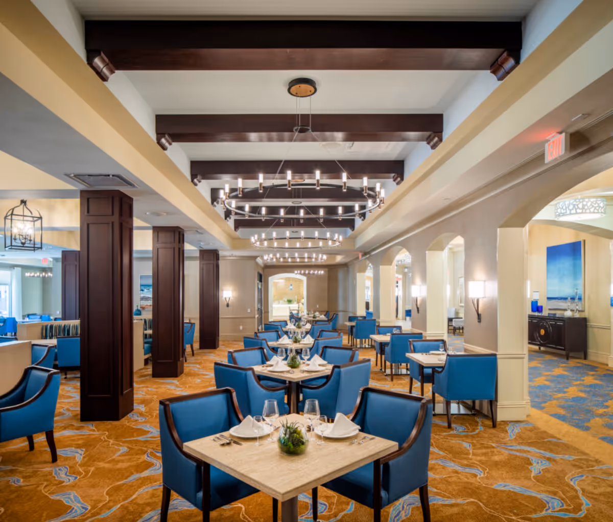 A spacious, elegant dining room with multiple square tables set with white napkins, glassware, and small green plants. The room features blue upholstered chairs, dark wooden beams on the ceiling, modern circular chandeliers, and a patterned carpet in shades of orange and blue. The walls have arches and sconces, and there is artwork and additional seating visible in the background.