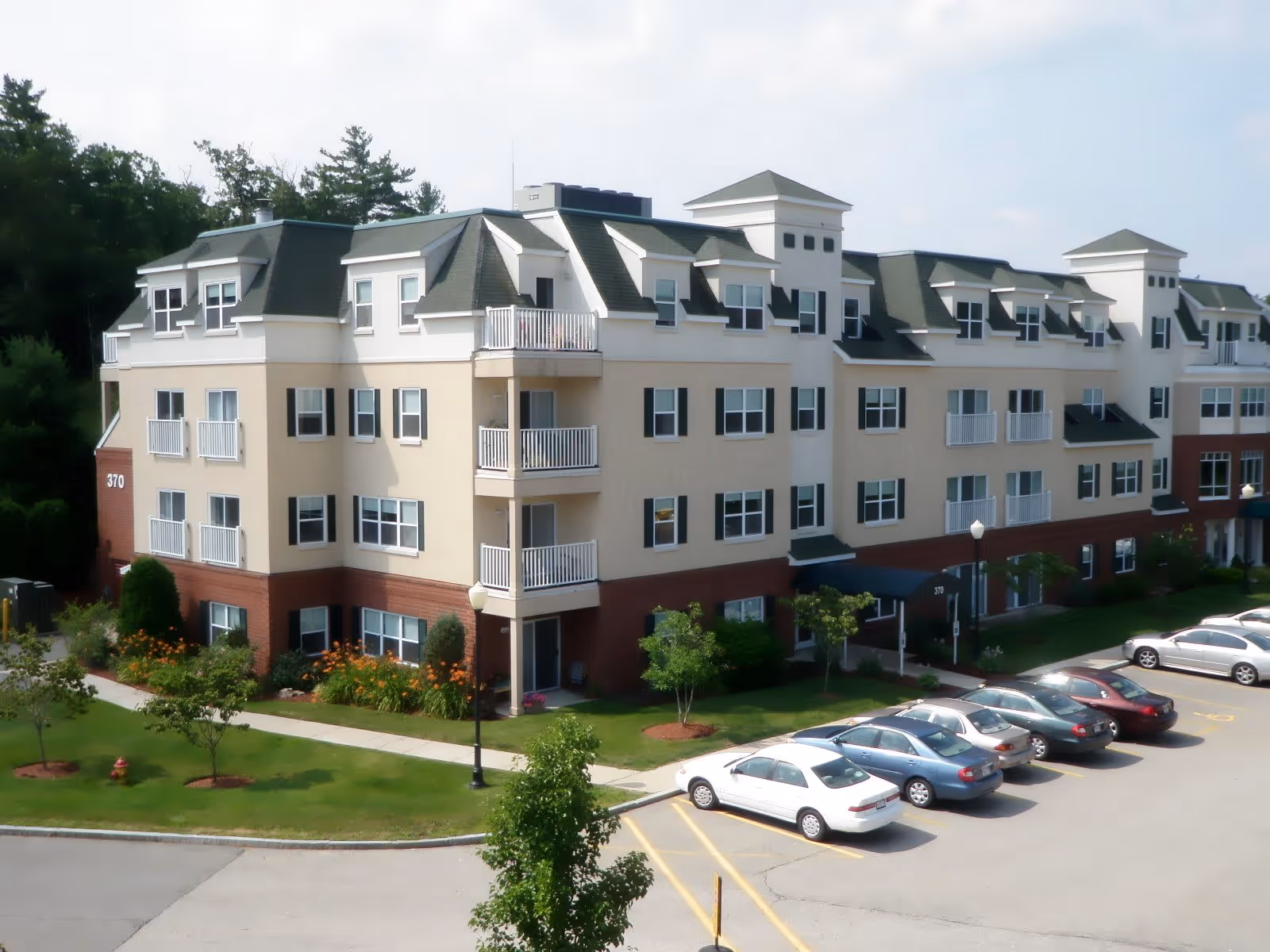 Exterior view of a multi-story senior living facility building with beige and brick facade, multiple windows, balconies, and a parking lot with several parked cars in front. The building is surrounded by green lawns, small trees, and flower beds.