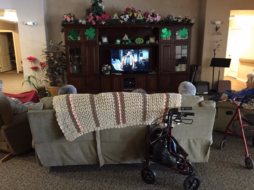 Residents seated on sofas watching a television in a communal living room with a wooden entertainment center and walkers in the foreground.