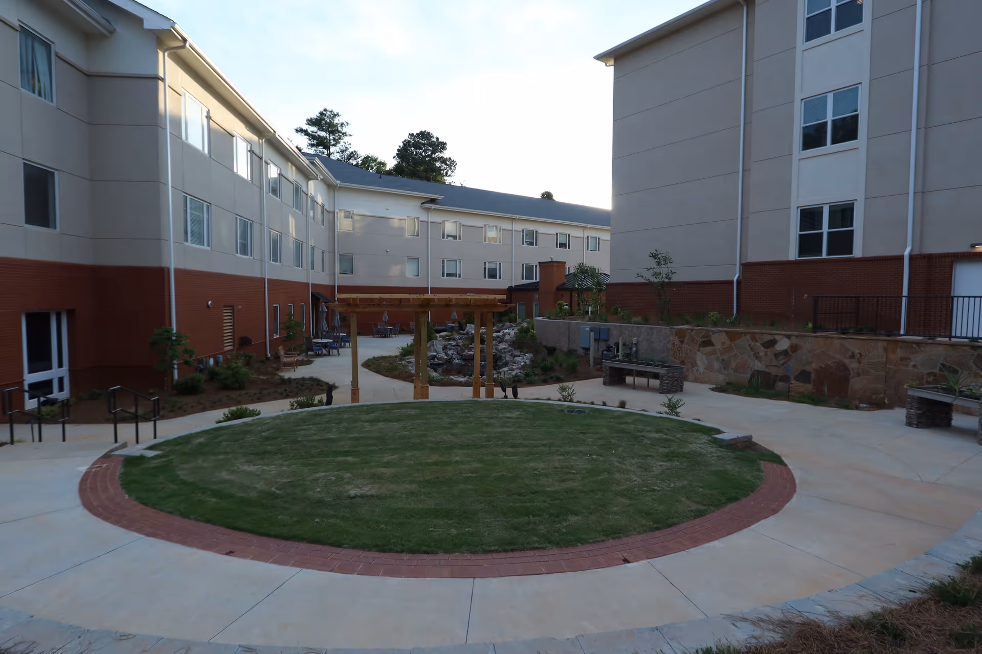 Outdoor courtyard area of a senior living facility with a circular grass patch surrounded by a concrete walkway. The courtyard is bordered by multi-story buildings with windows, and there is a wooden pergola structure over a pathway leading to a small rock waterfall feature. Benches and plants are placed around the area.