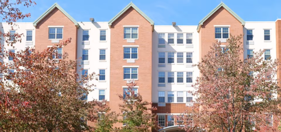 Front exterior of a multi-story brick-and-white senior living building with gabled rooflines, rows of windows, and trees with autumn foliage in front.