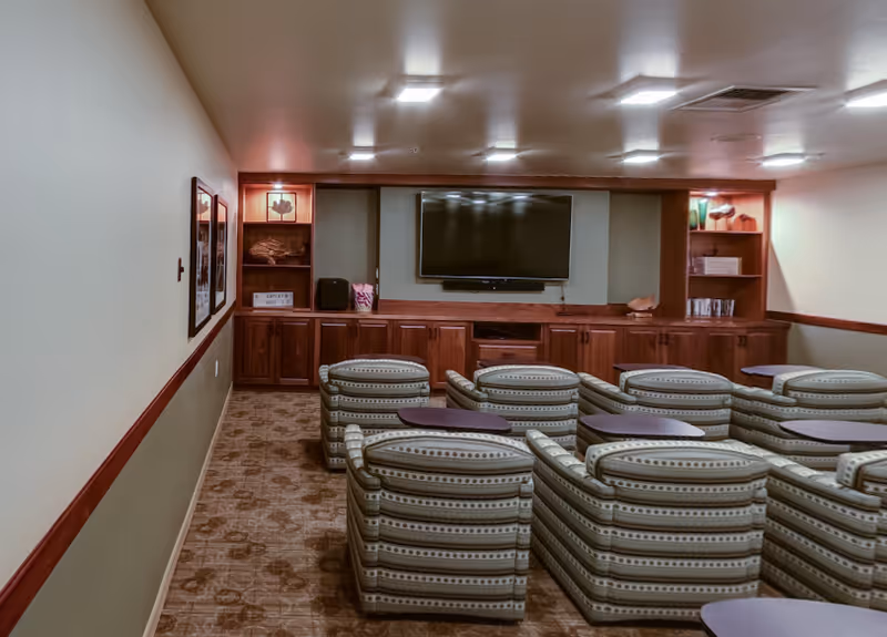 A small theater room with two rows of patterned upholstered chairs facing a large flat-screen TV mounted on a wooden cabinet wall. The room has beige walls, carpeted floor, and built-in shelves with decorative items on either side of the TV.