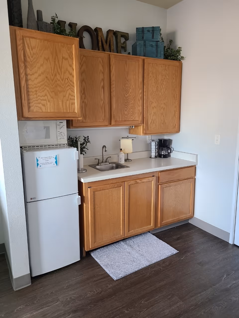 A small kitchen area with wooden cabinets, a white countertop, a small sink, a coffee maker, a paper towel holder, and a small white refrigerator with a microwave on top. Decorative items including plants, blue storage boxes, and large letters spelling HOME are placed on top of the cabinets. A gray rug is on the dark wood floor in front of the sink.