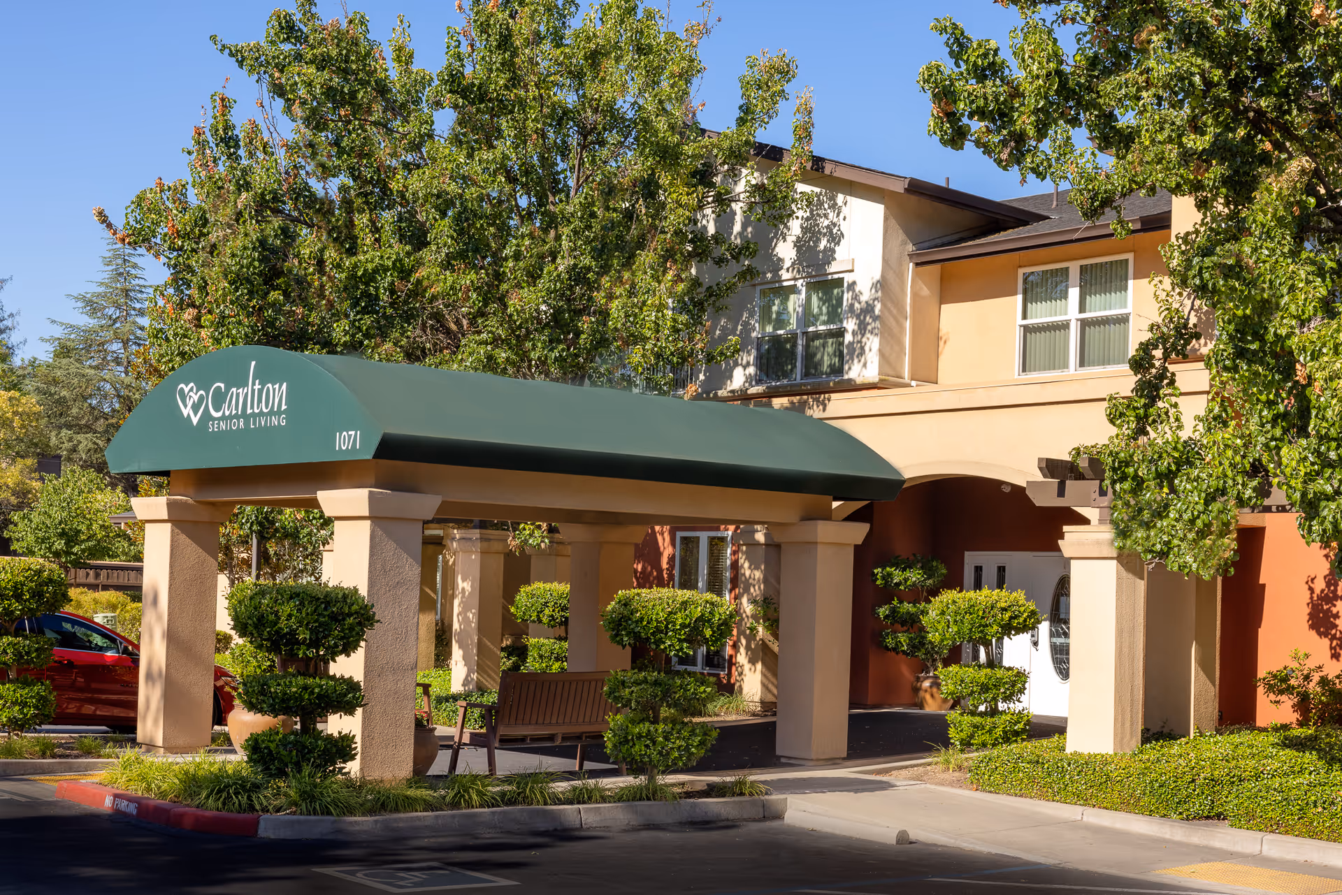 Entrance to Carlton Senior Living Sacramento Atrium Building with a green canopy displaying the facility name and number 1071, surrounded by neatly trimmed bushes and trees under a clear blue sky.