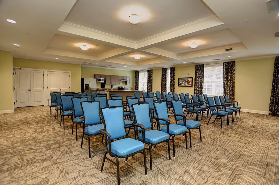 A spacious room with rows of blue cushioned chairs arranged facing forward. The room has a beige carpet, light green walls, and a ceiling with recessed lighting. At the back of the room, there is a kitchenette area with cabinets, a refrigerator, microwave, and stove. Windows with dark patterned curtains allow natural light into the room.