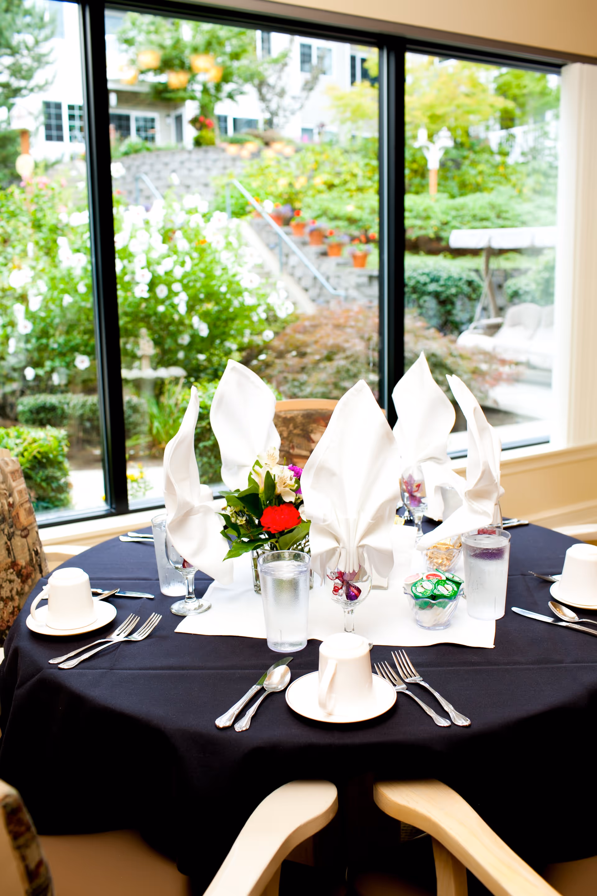 A round dining table set with folded white napkins, cups, glasses, silverware, and a floral centerpiece in front of a large window overlooking a garden.