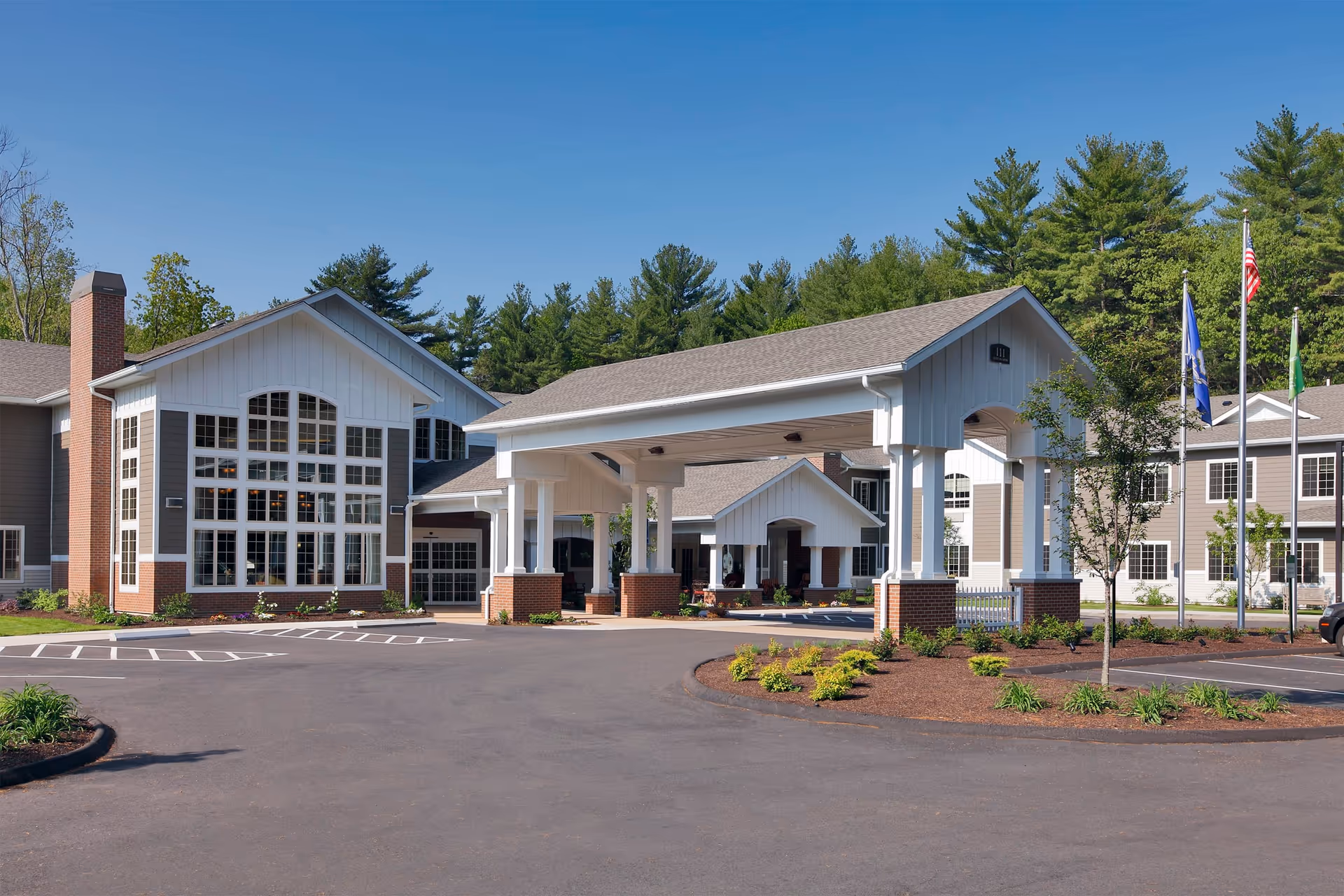 Front entrance of a senior living facility with a covered porte-cochère, large windows, flags, and landscaped driveway.