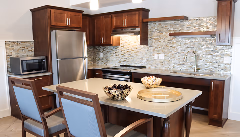 Modern kitchen with dark wood cabinets, a stainless steel refrigerator, microwave, stove, and a sink with a faucet. There is a kitchen island with a beige countertop, two wooden chairs with blue cushions, and two decorative bowls and a round tray on the island. The backsplash is made of small rectangular tiles in neutral tones.