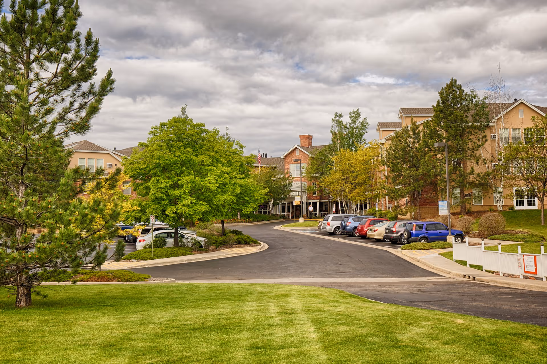 View of the Someren Glen Retirement Community showing a paved driveway lined with parked cars on the right side, surrounded by green trees and well-maintained grass under a cloudy sky. The multi-story residential building is visible in the background.