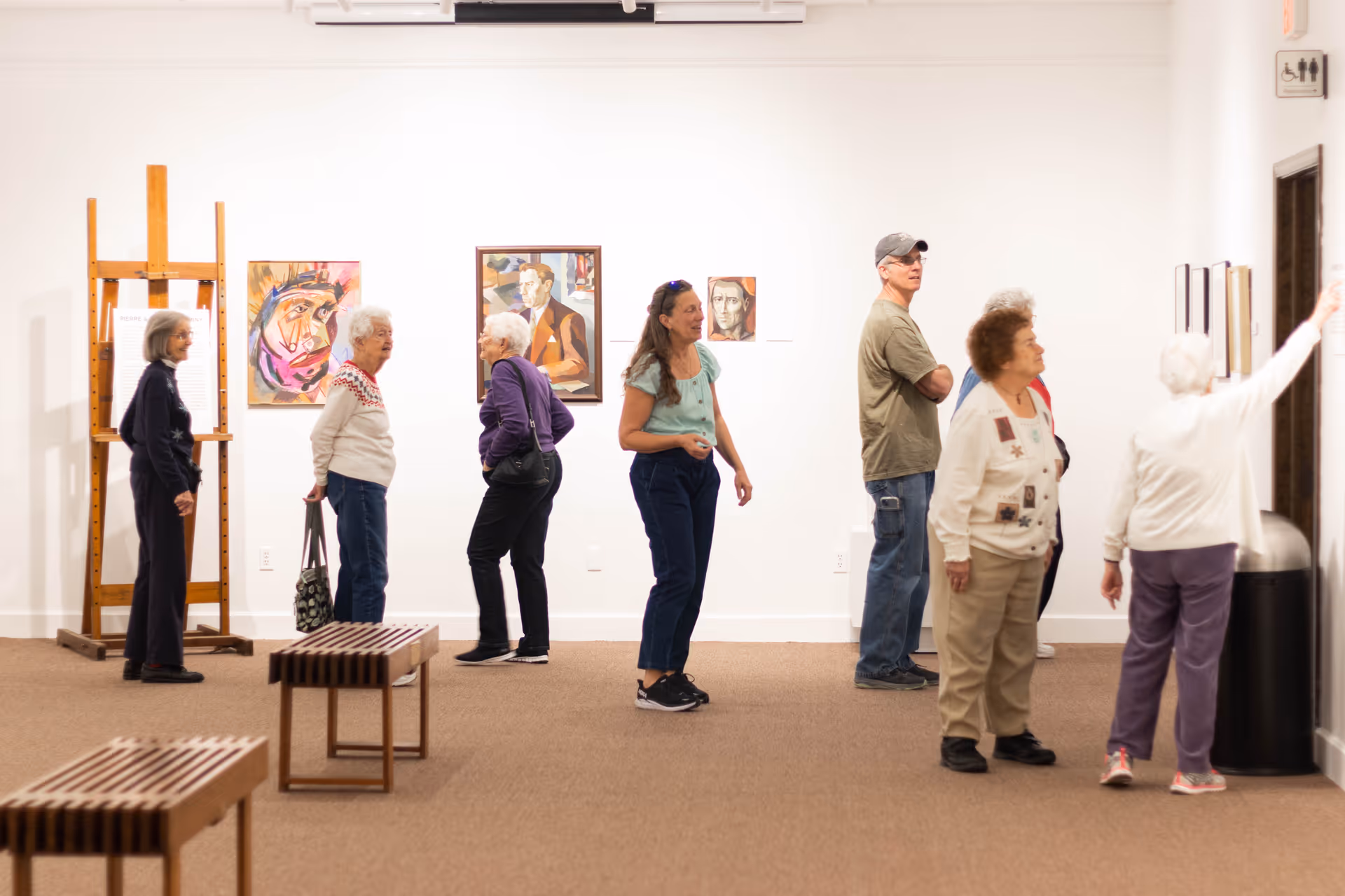 A group of elderly people and one younger adult standing and observing artwork displayed on the walls of a gallery-like room with white walls and brown carpet. There are wooden benches in the foreground and an easel on the left side.