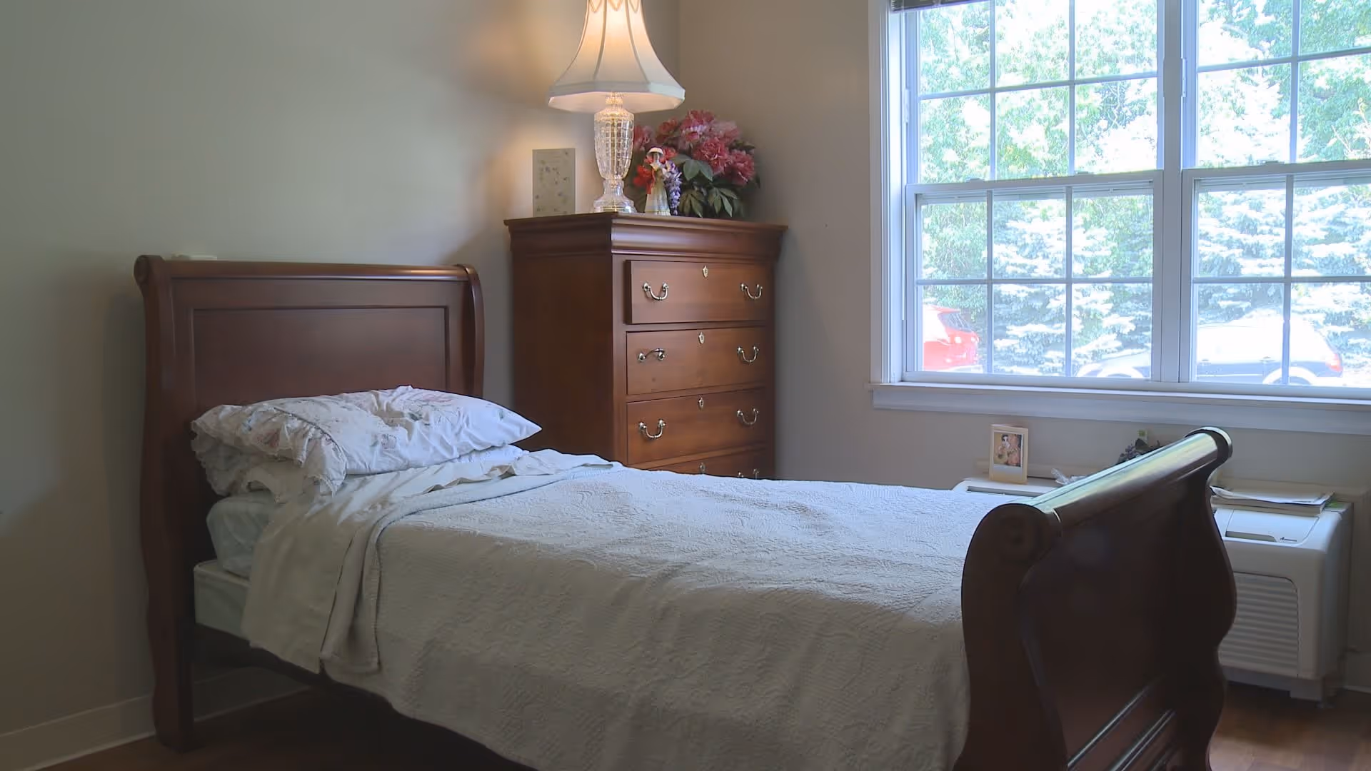 A neatly made single bed with white bedding and pillows in a senior living bedroom. Next to the bed is a wooden chest of drawers with a decorative lamp and a flower arrangement on top. A large window lets in natural light, showing greenery and parked cars outside.