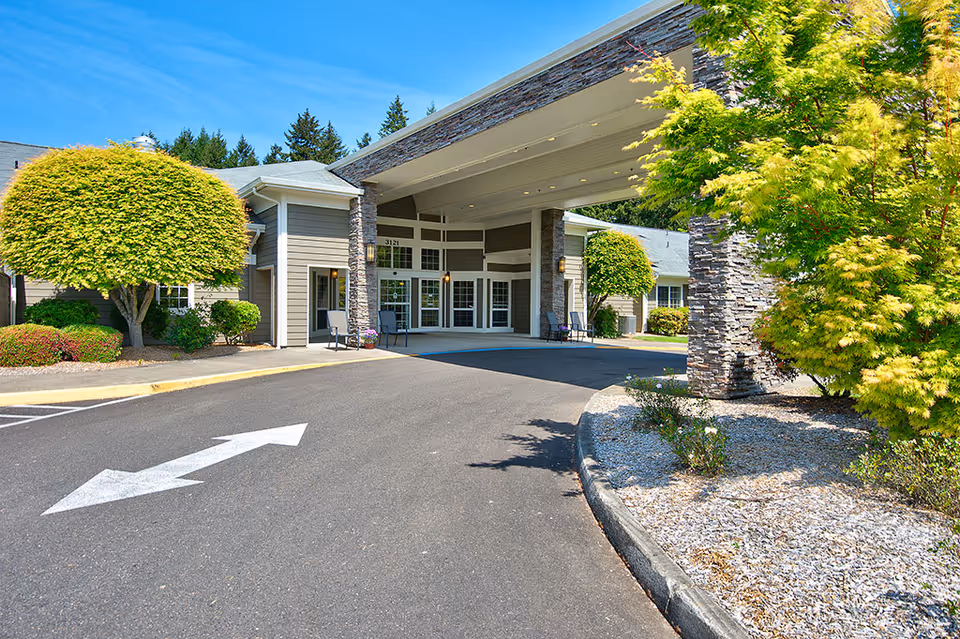 Covered porte-cochère entrance and drive-up area of a retirement community building with manicured trees and shrubs.