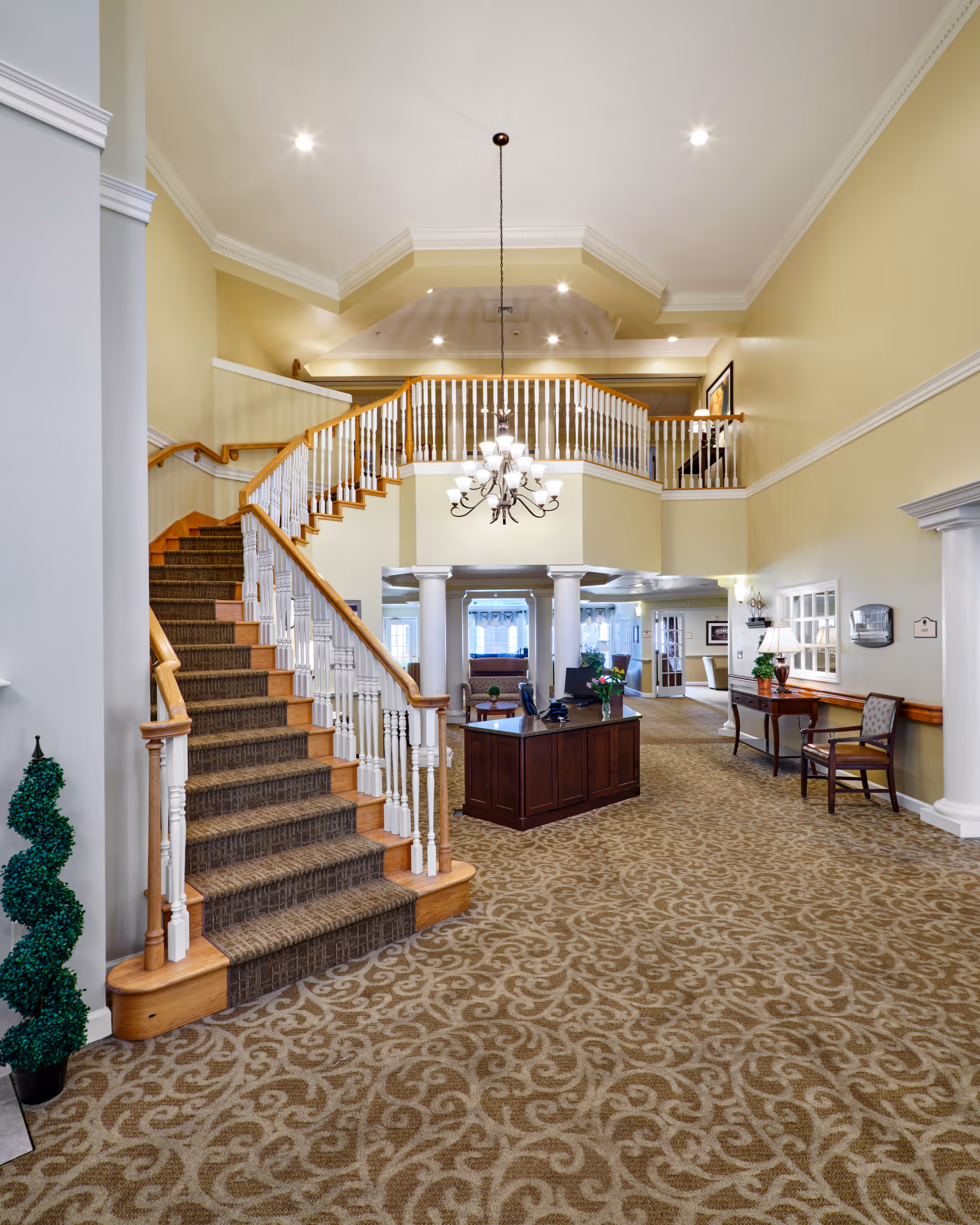 Spacious assisted living lobby with a carpeted grand staircase, chandelier, reception desk, and seating area.