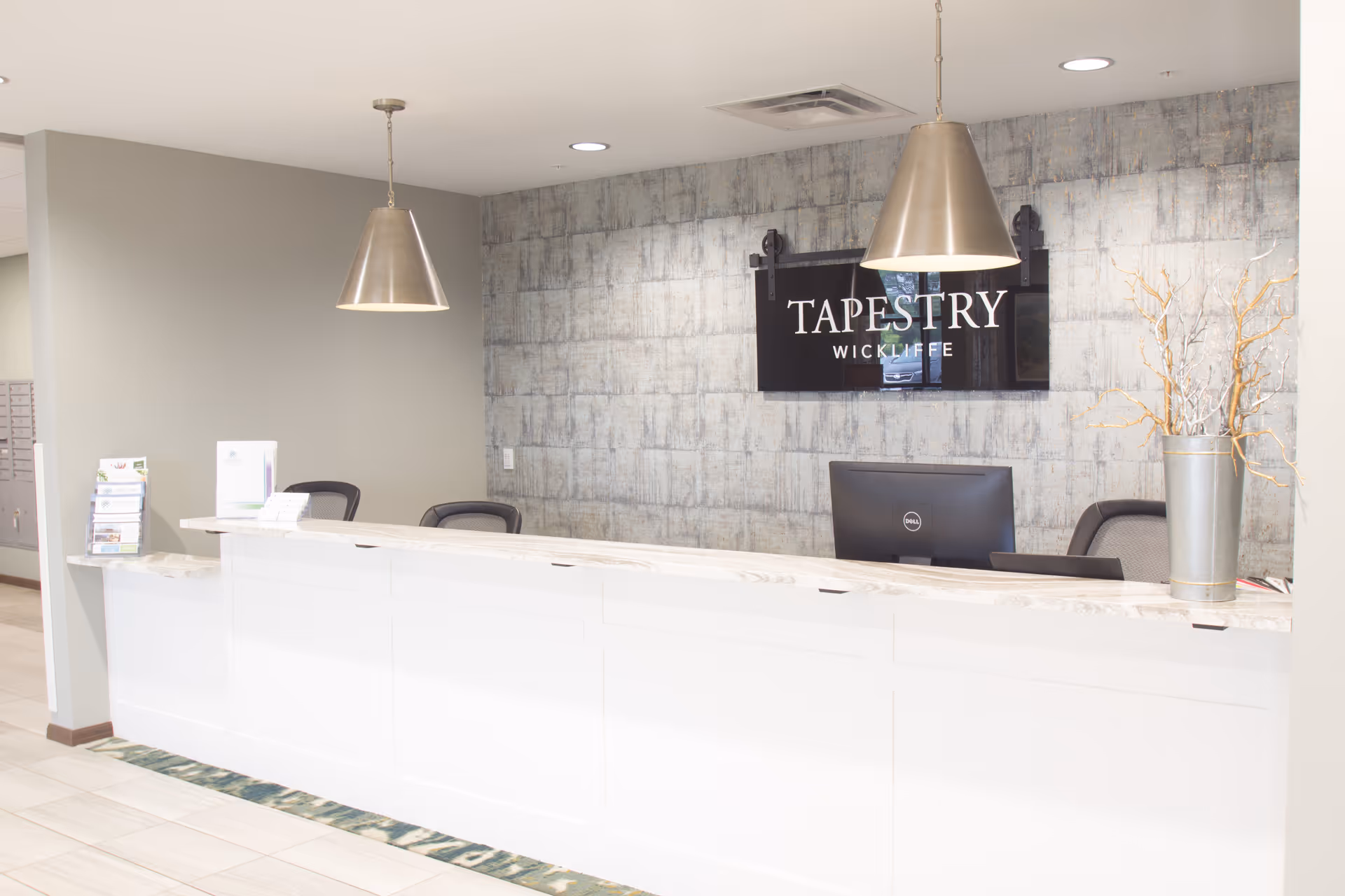 Bright reception desk with a white counter, two pendant lights, chairs, and a wall sign reading "TAPESTRY WICKLIFFE".