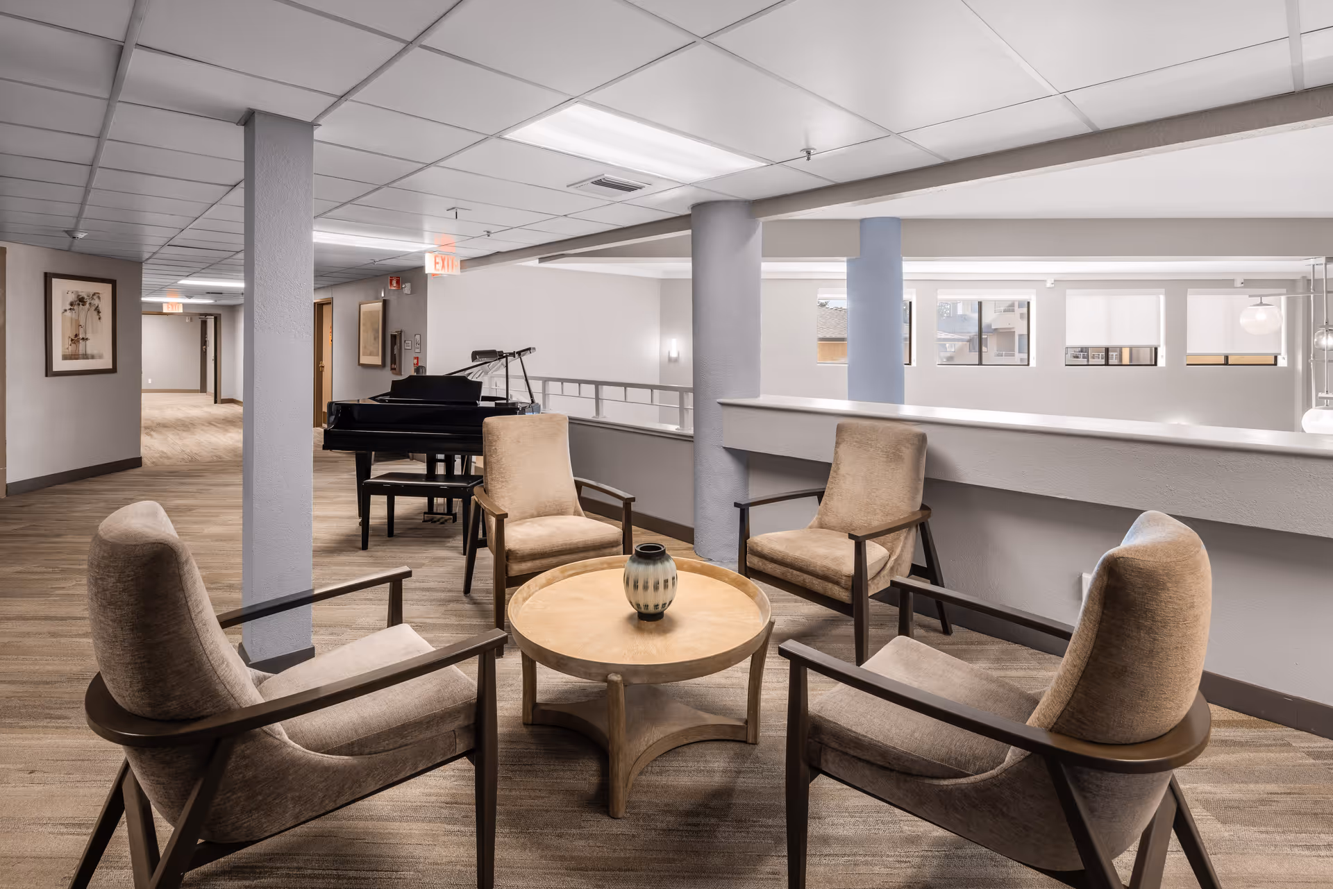 A cozy seating area in a senior living community with four cushioned armchairs arranged around a round wooden table with a decorative vase on top. In the background, there is a black grand piano and a hallway with framed artwork on the walls. The space has a neutral color palette with beige and gray tones and large windows letting in natural light.