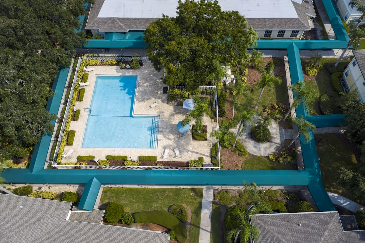 Aerial view of a senior living facility courtyard featuring a rectangular swimming pool with lounge chairs around it, surrounded by a fence and landscaped greenery including trees and bushes. The courtyard is enclosed by buildings with pathways and additional greenery.