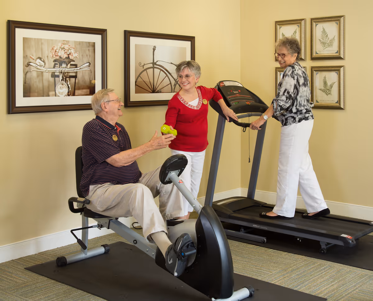 Three elderly individuals in a fitness room at a senior living facility. One man is seated on a recumbent exercise bike, smiling and reaching out to a woman in a red top who is handing him a small green dumbbell. Another woman is walking on a treadmill, smiling and looking at the other two. The room has beige walls with framed pictures and a carpeted floor with exercise mats under the equipment.