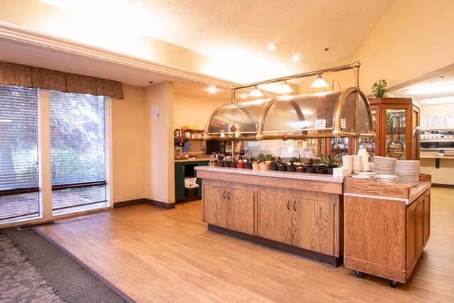 Interior view of a dining area in a senior living facility featuring a wooden buffet counter with a glass sneeze guard, plates stacked on the side, and various food items underneath. Large windows with blinds allow natural light to enter, and there is a cabinet and coffee station in the background.