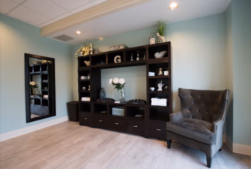 A cozy interior room with light blue walls and light wood flooring. There is a large dark wood shelving unit filled with decorative items, towels, and a vase with white flowers in the center. To the right of the shelving unit is a plush gray armchair. On the left wall, there is a tall rectangular mirror with a dark frame.
