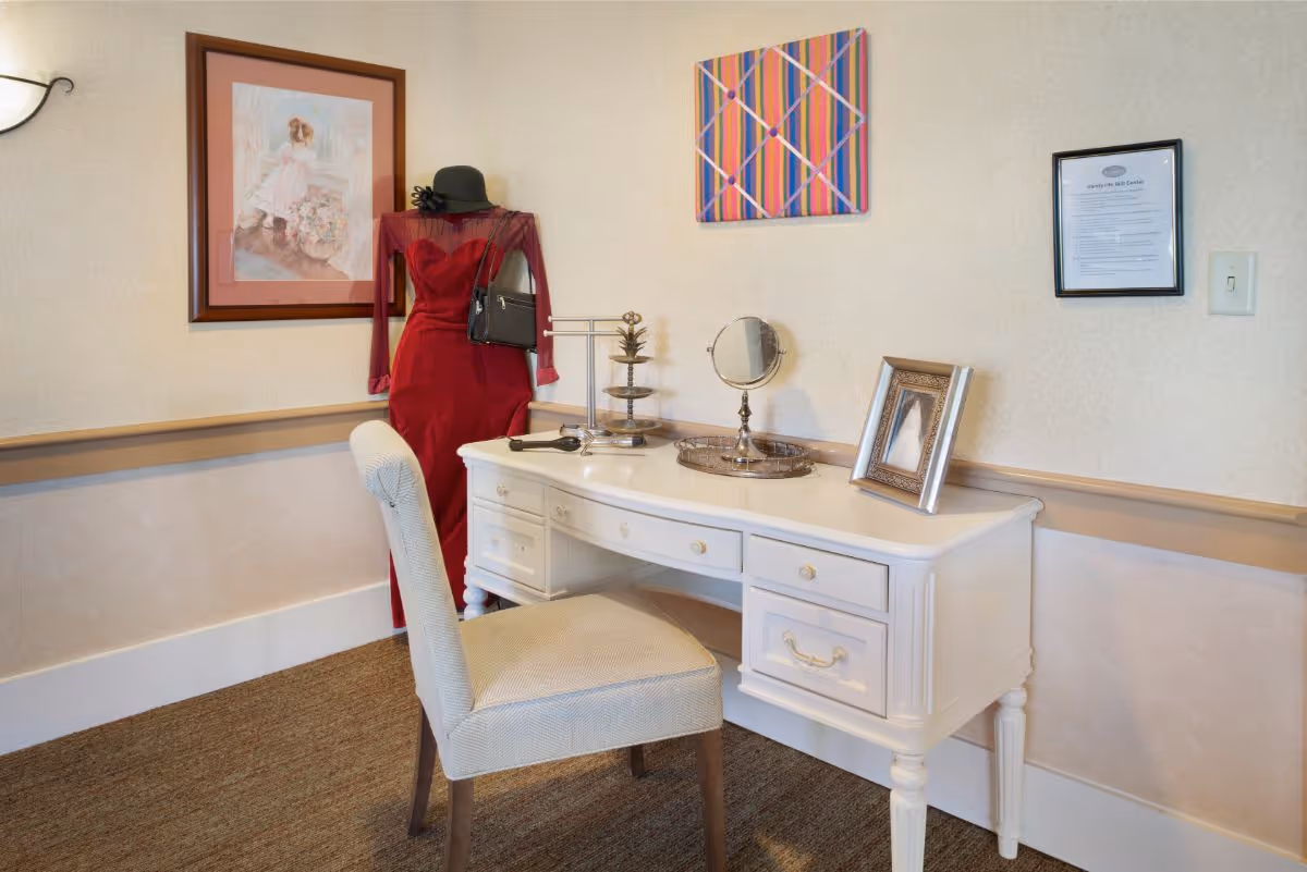 A white vanity desk with drawers and a cushioned chair in front of it. On the desk are a small round mirror, a framed photo, a tiered tray, and a hairbrush. To the left of the desk is a mannequin dressed in a red dress, black hat, and black handbag. The walls have framed artwork and a document in a black frame.