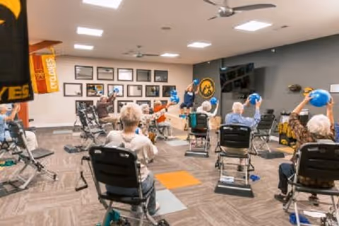 A group of elderly individuals seated in chairs in a spacious room, participating in a fitness or exercise class using blue exercise balls. The room has carpeted flooring, ceiling fans, framed pictures on the wall, and a large TV screen.
